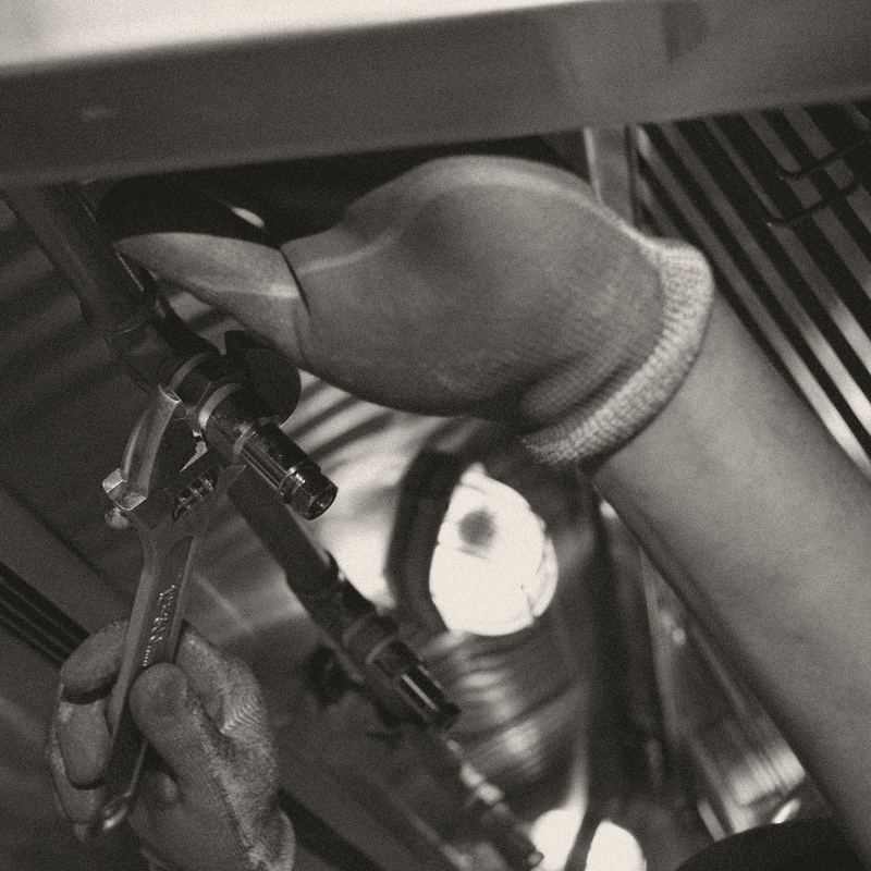 Close-up of a gloved hand holding an adjustable spanner on a pipe fitting inside a steel kitchen hood.