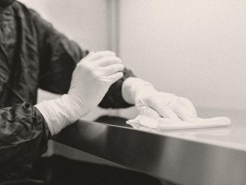 Two white-gloved hands wiping a stainless steel work surface with a cloth.