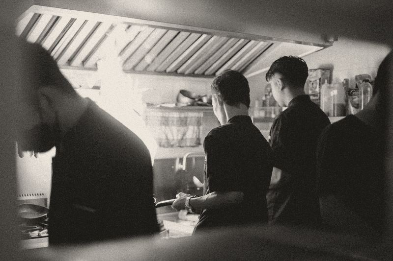 Chefs at a busy cooking line with a wok flame rising towards the stainless steel hood above.